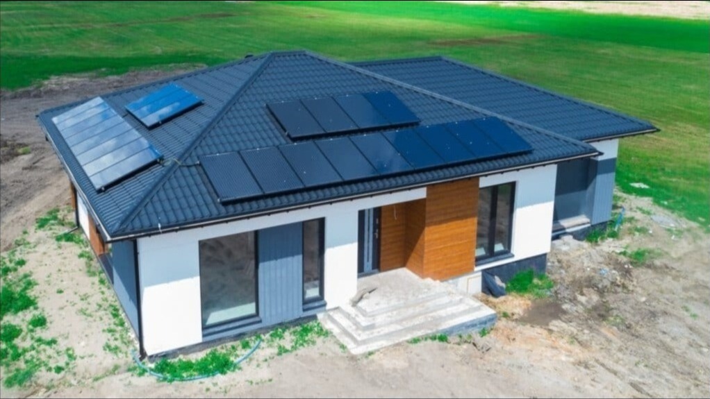 Nowoczesny dom jednorodzinny z ciemną dachówką i panelami fotowoltaicznymi w dwóch różnych konfiguracjach: panele standardowe i panele z odbijającą powierzchnią, na tle zielonego trawnika.