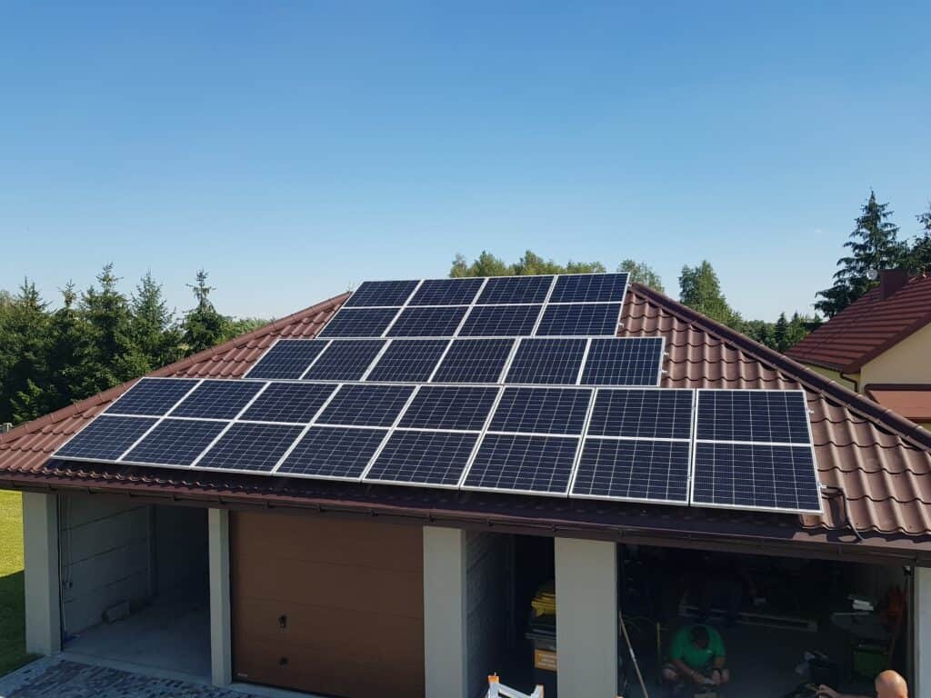 Instalacja paneli słonecznych na dachu garażu z brązową dachówką, widok z góry, błękitne niebo w tle.