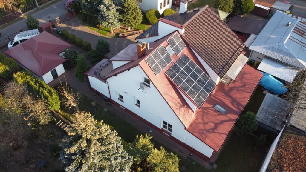 Widok z drona na dom jednorodzinny z panelami fotowoltaicznymi na dachu o czerwonej dachówce, otoczony zielenią i innymi budynkami.