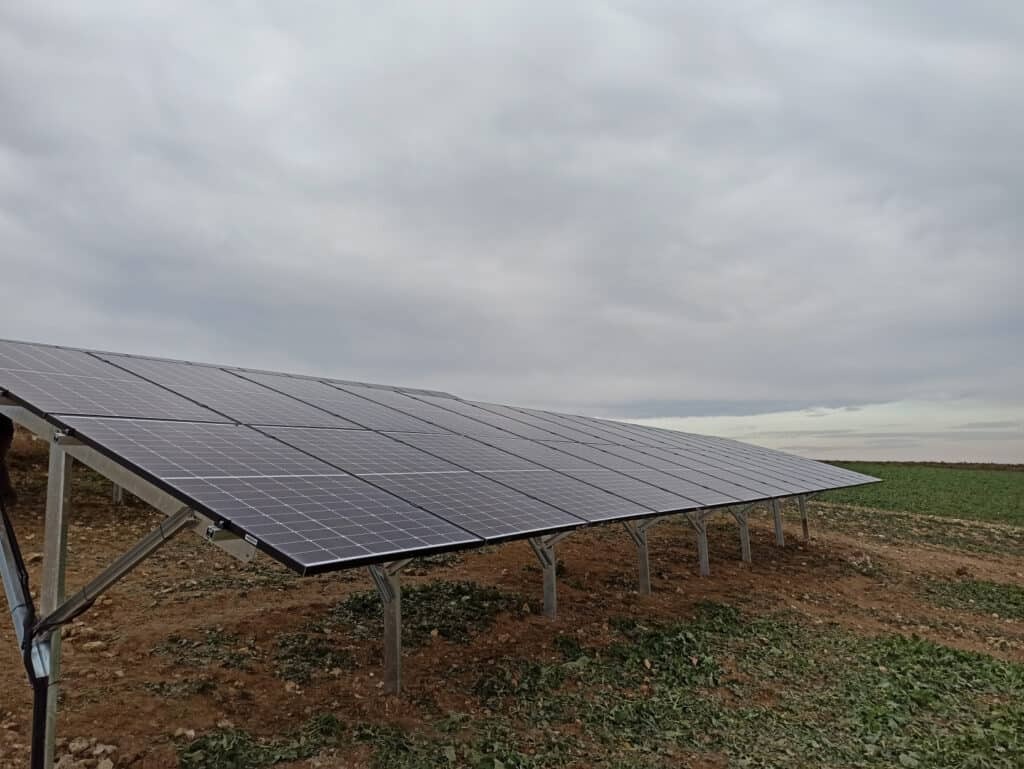 Rząd paneli słonecznych zamontowanych na metalowej konstrukcji na polu, pochmurne niebo w tle.