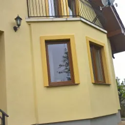 Żółta elewacja domu jednorodzinnego z balkonem i brązowymi ramami okiennymi, widoczne schody wejściowe i fragment chodnika z kostki brukowej, na balkonie antena satelitarna.