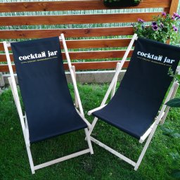 Seleo Druk - Dwa drewniane leżaki z czarnym płótnem z napisem 'Cocktail Jar - Usługi Barmańskie' stoją na trawie przed drewnianym płotem z fioletowymi kwiatami w donicach.