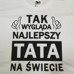 Biała koszulka z czarnym nadrukiem 'Tak wygląda najlepszy tata na świecie' oraz grafiką dwóch dłoni z podniesionymi kciukami, leżąca na szarym tle.