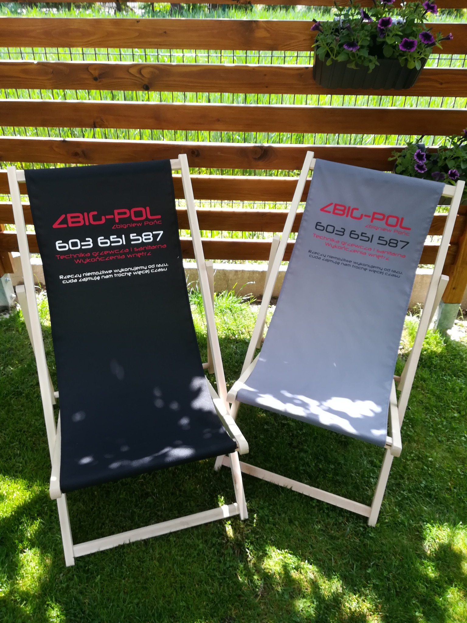 Dwa leżaki plażowe z nadrukowanym logo firmy ZBIC-POL, jeden w kolorze czarnym, drugi szary, stoją na trawie przed drewnianym ogrodzeniem z kwiatami.