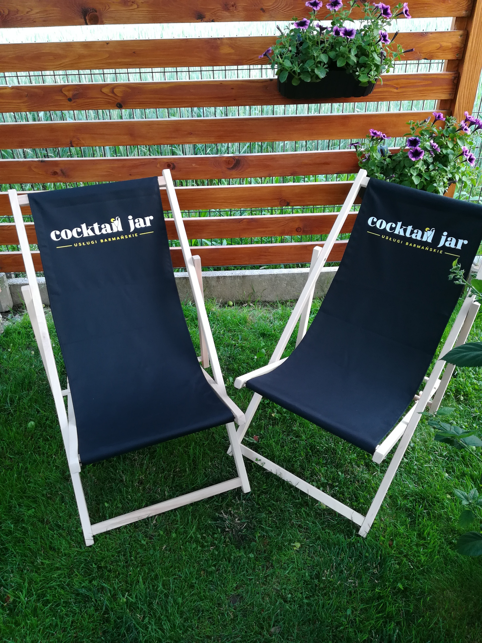 Dwa drewniane leżaki z czarnym płótnem z napisem 'Cocktail Jar - Usługi Barmańskie' stoją na trawie przed drewnianym płotem z fioletowymi kwiatami w donicach.