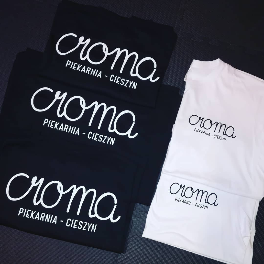 Stos czarnych i białych koszulek z białym napisem 'croma PIEKARNIA - CIESZYN', ułożonych na ciemnym tle.