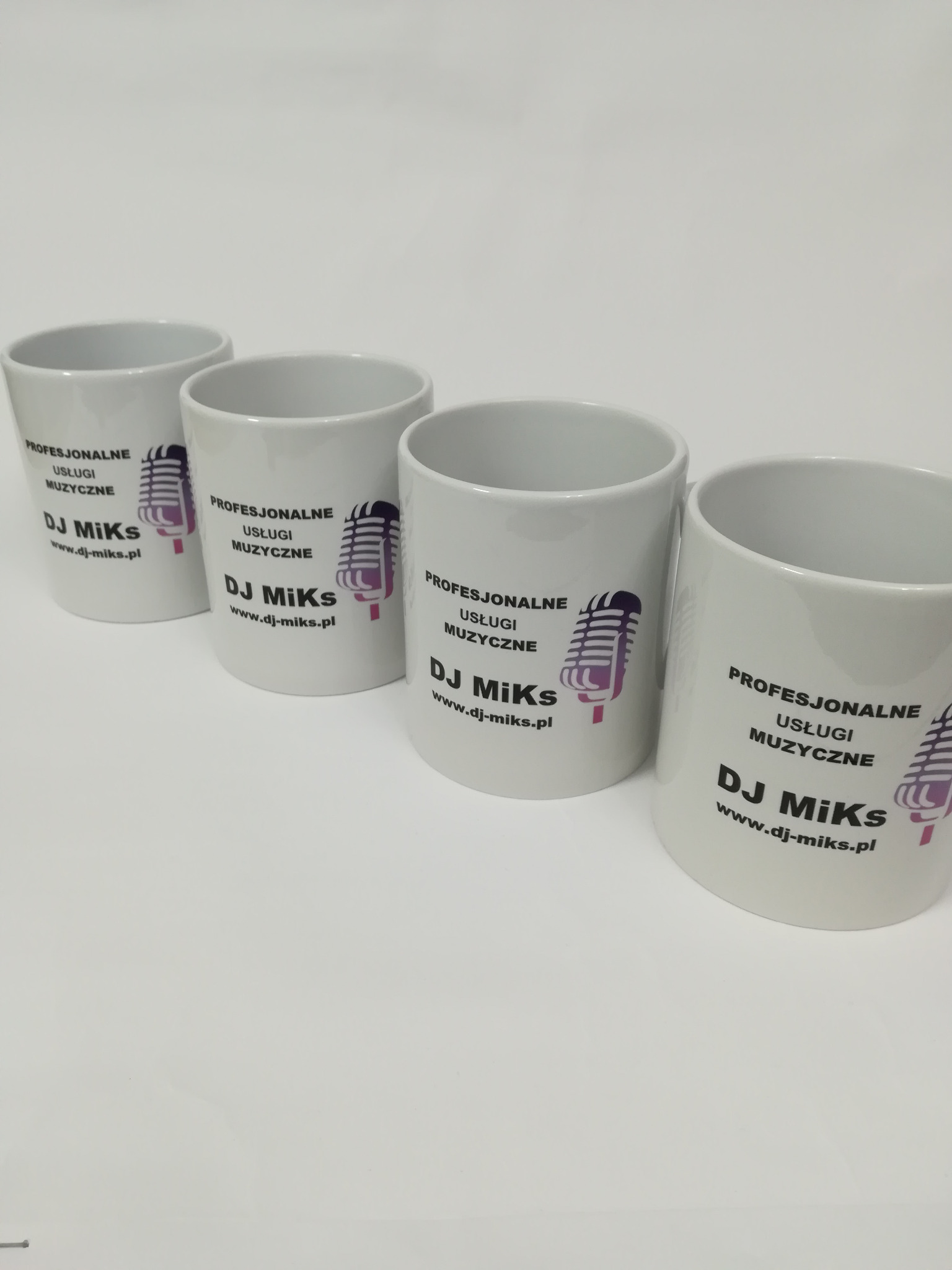 Cztery białe kubki z nadrukiem logo DJ Miks oraz grafiką mikrofonu w odcieniach fioletu, ustawione w rzędzie na białym tle.