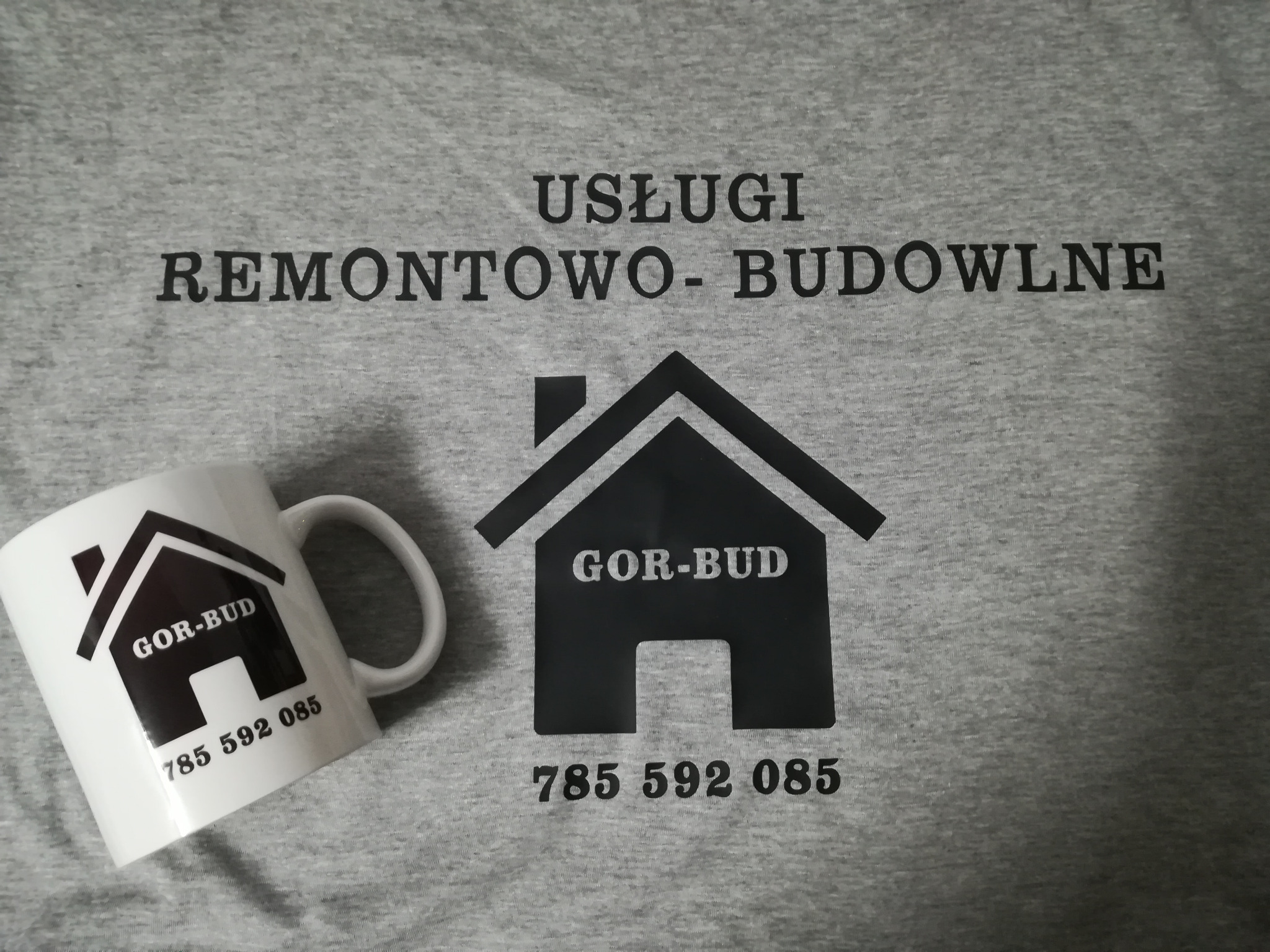 Szara koszulka z nadrukiem 'USŁUGI REMONTOWO-BUDOWLNE', logo firmy w kształcie domu 'GOR-BUD' i numerem telefonu, obok biały kubek z identycznym nadrukiem, leżący pod kątem.