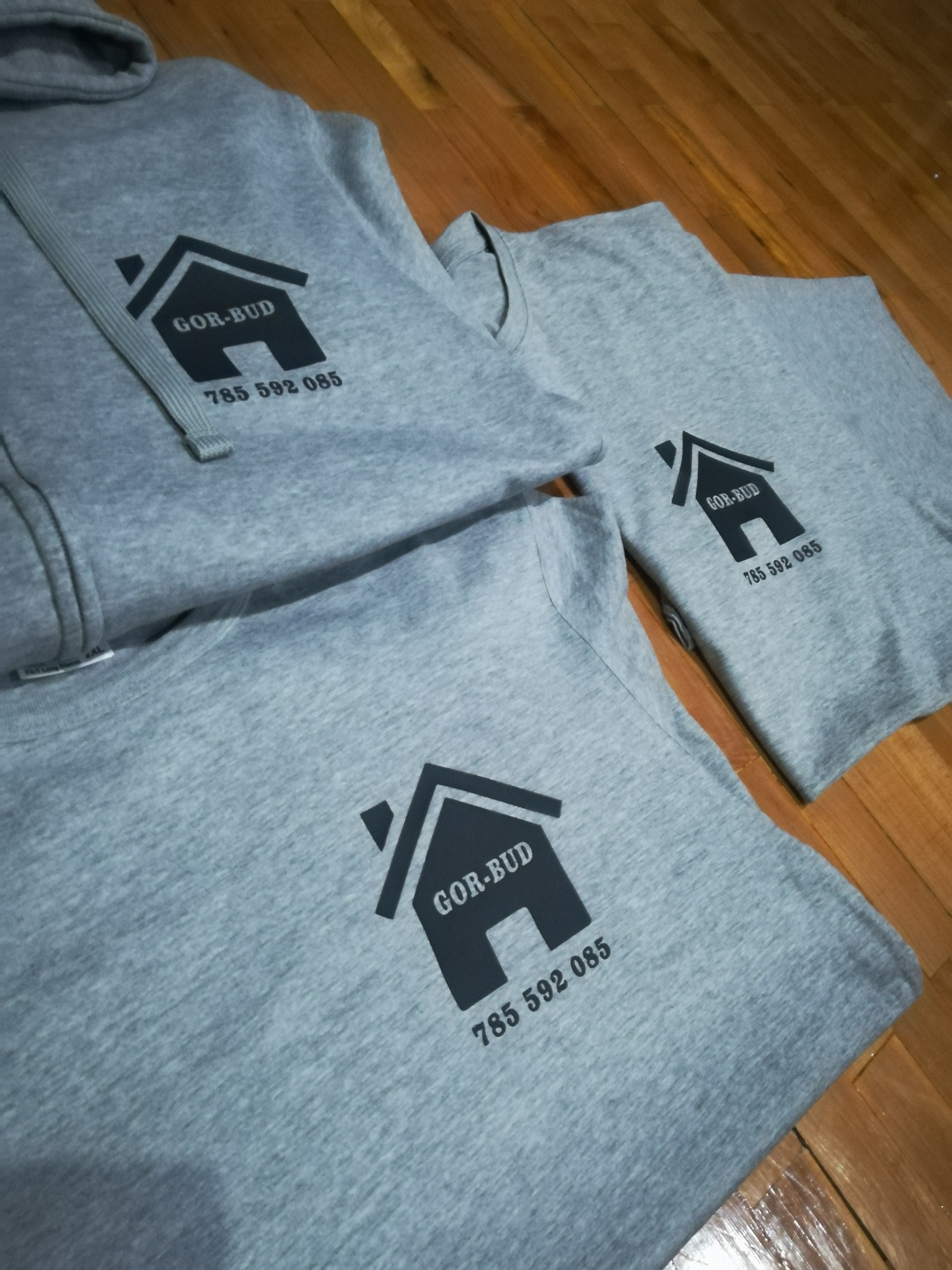 Trzy szare ubrania - bluza z kapturem i dwie koszulki - z czarnym nadrukiem logo firmy GOR-BUD i numerem telefonu, ułożone na drewnianej podłodze.