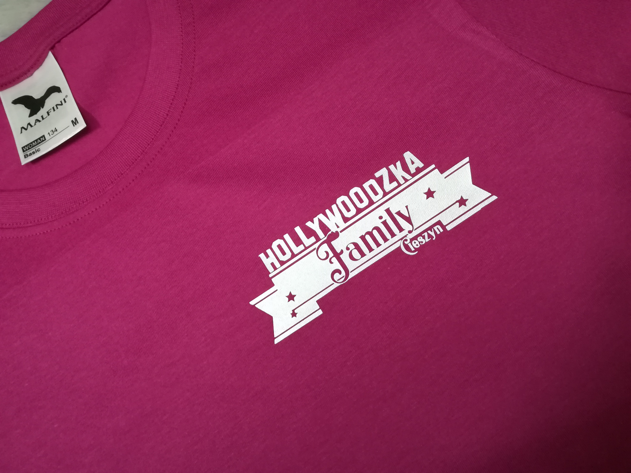 Malinowa koszulka damska z białym nadrukiem 'Hollywoodzka Family Cieszyn', widoczna metka producenta Malfini.