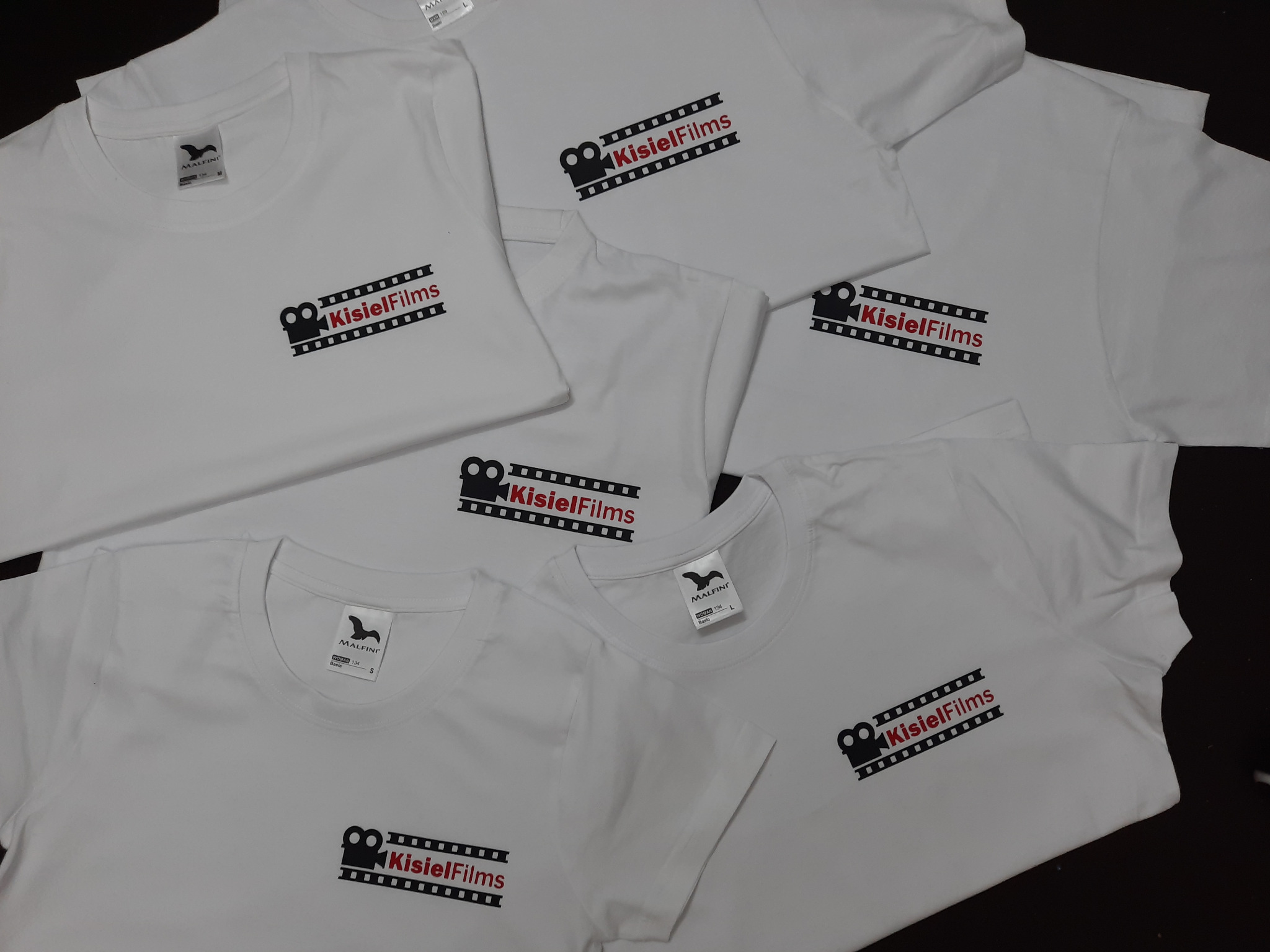 Stos białych koszulek z nadrukowanym logo 'KisielFilms' w formie kamery filmowej i napisu na tle imitującym taśmę filmową.