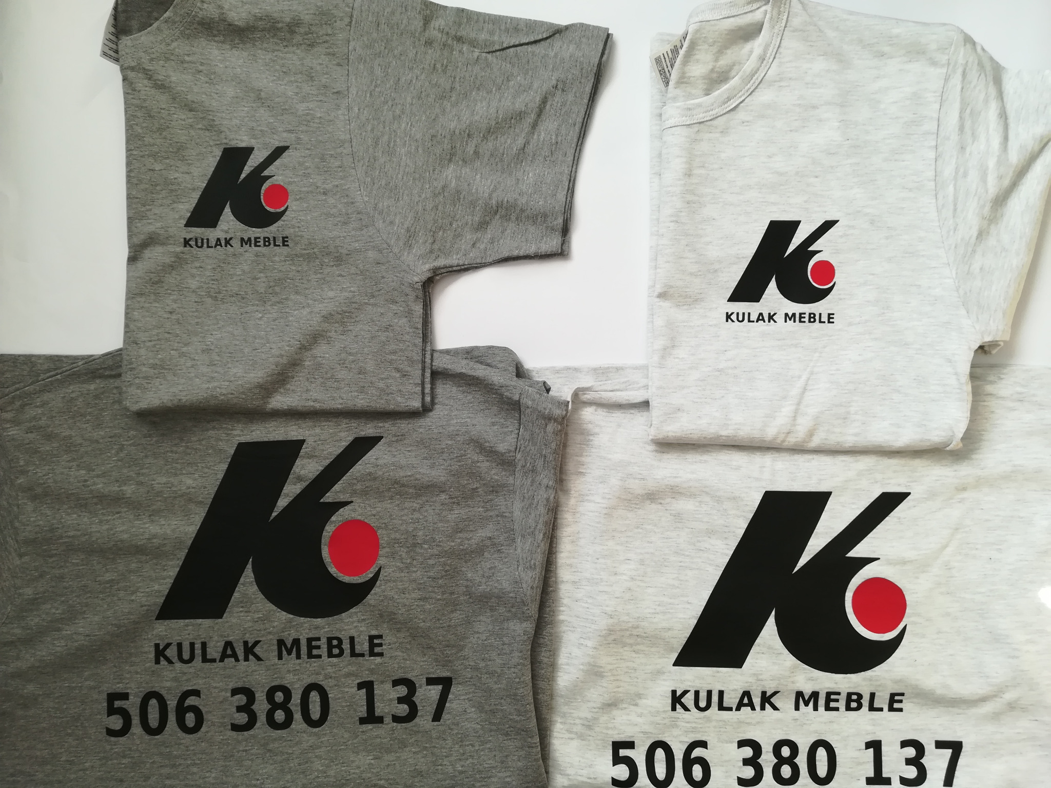 Dwa szare i dwa białe t-shirty z nadrukowanym logo 'KULAK MEBLE' oraz numerem telefonu, ułożone na białym tle.