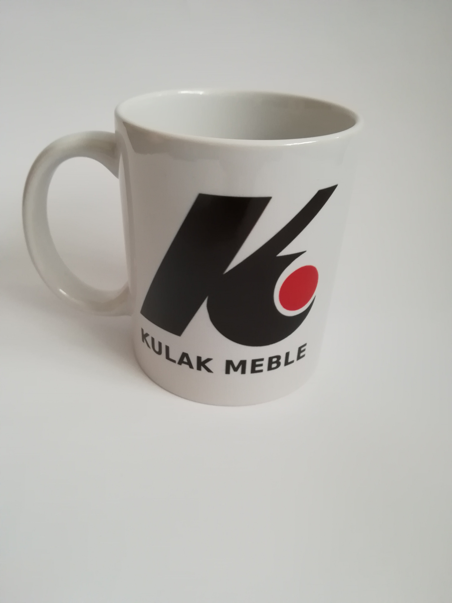 Biały kubek z czarno-czerwonym logo firmy Kulak Meble na jednolitym, jasnym tle.