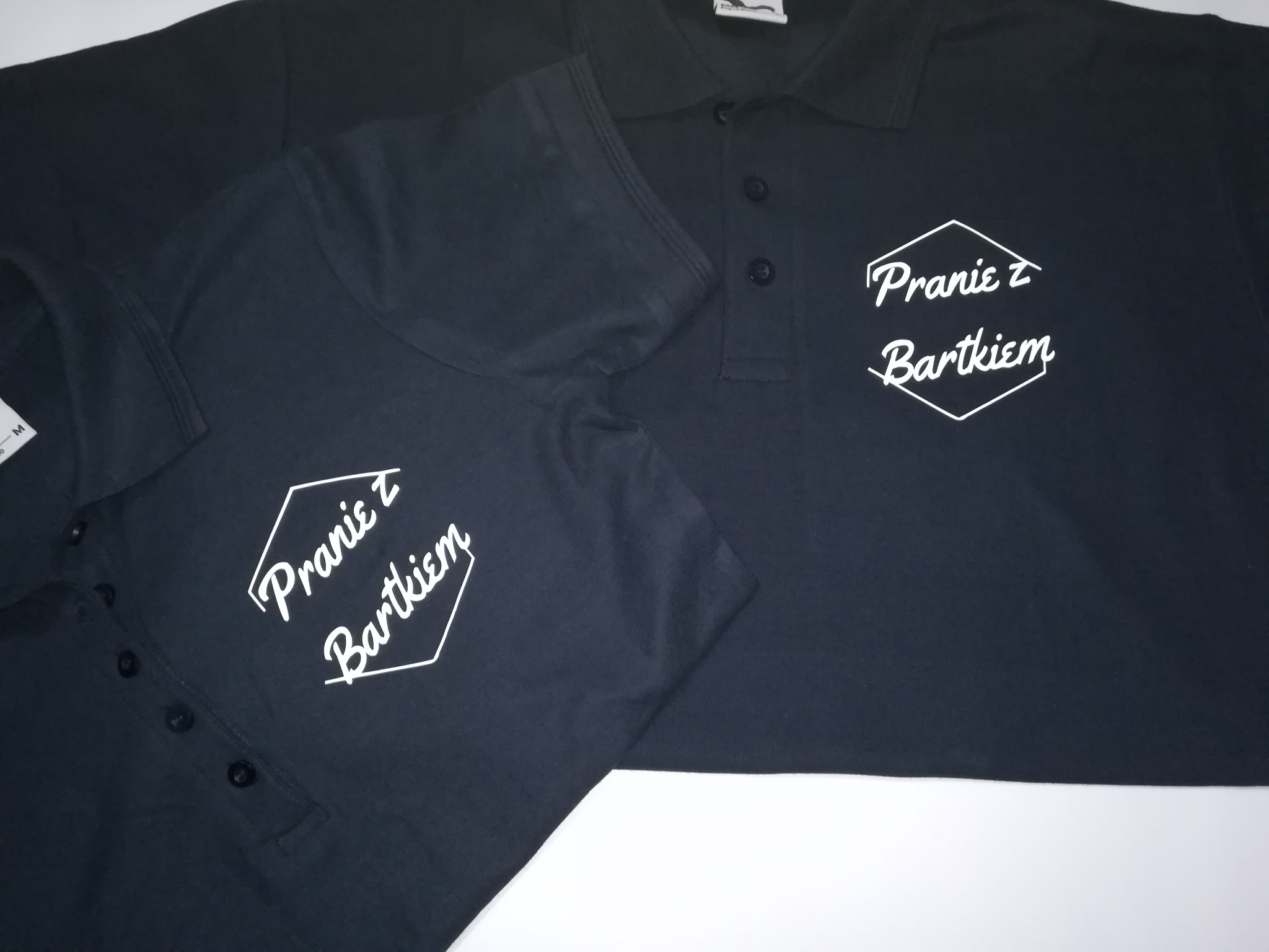Dwie czarne koszulki polo z białym nadrukiem 'Pranie z Bartkiem' w heksagonalnej ramce, leżące na białym tle.