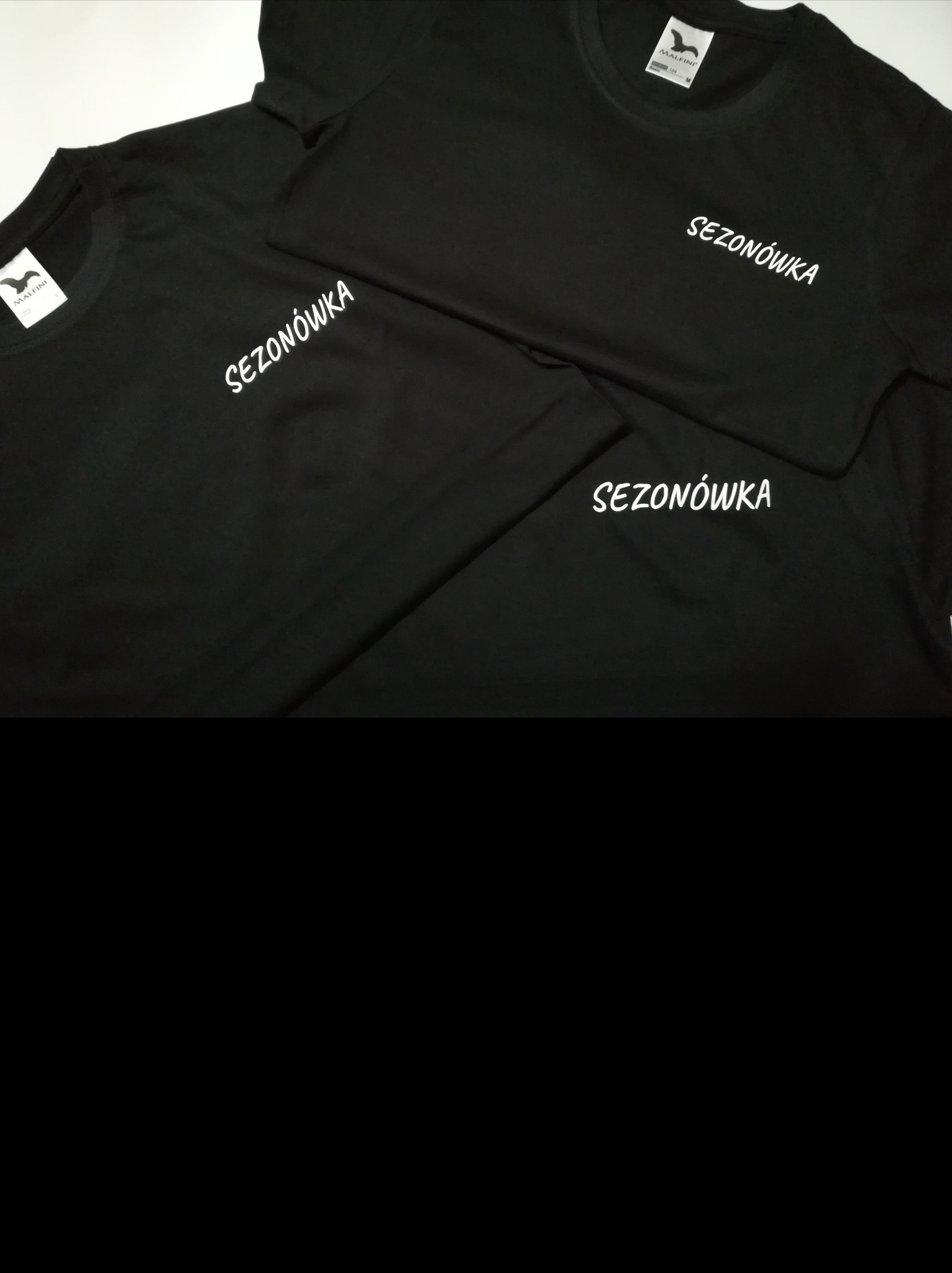 Trzy czarne koszulki z białym nadrukiem 'SEZONÓWKA' leżące na białym tle, widoczne metki producenta MALFINI.