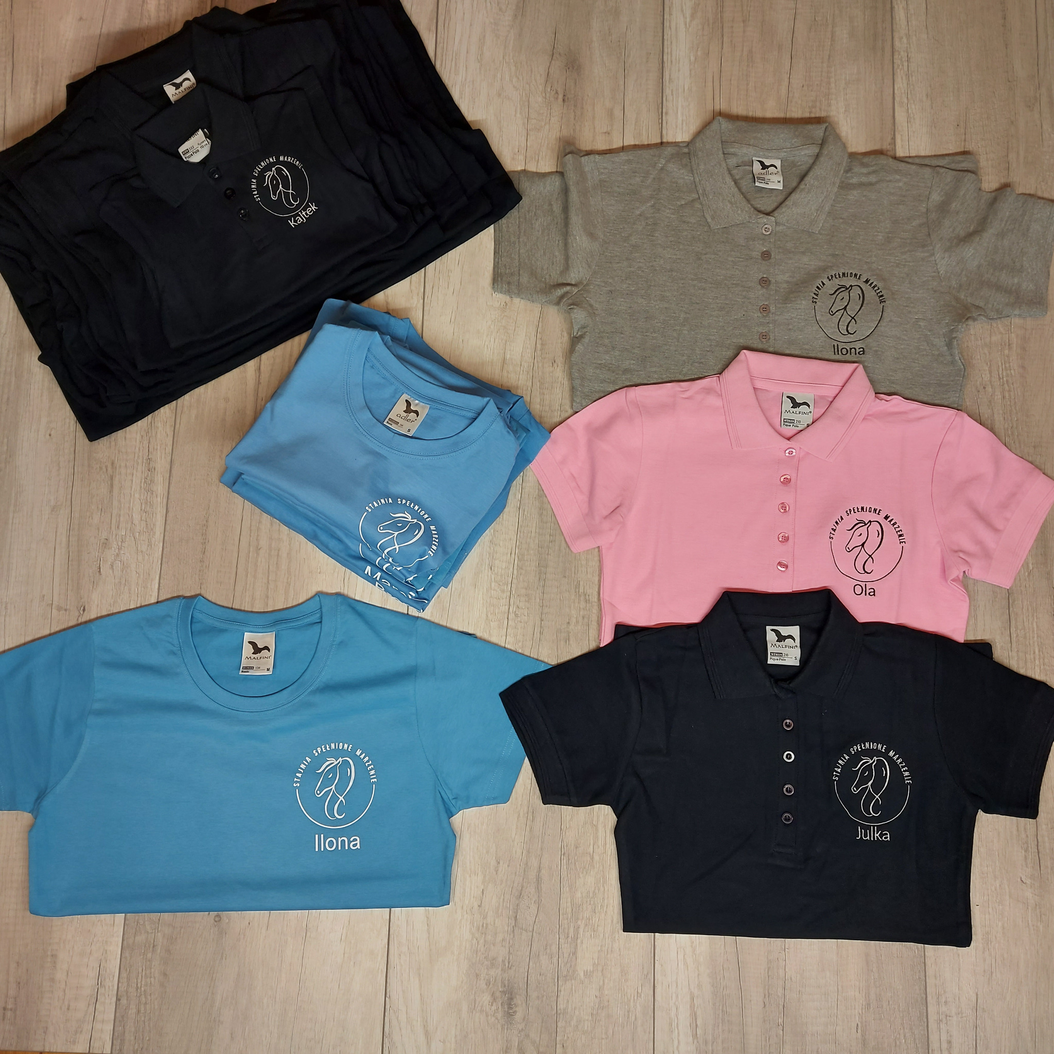 Różnokolorowe koszulki polo i t-shirty z nadrukowanym logo 'Stajnia Spełnione Marzenie' i imionami: Kajtek, Ilona, Ola, Julka, ułożone na drewnianym tle.