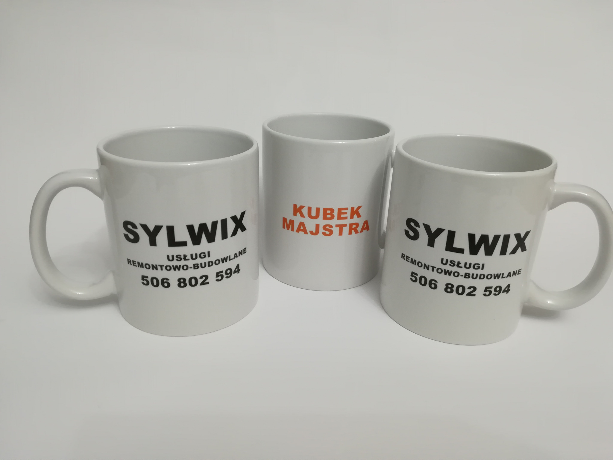 Trzy białe kubki z nadrukami: dwa z logo firmy remontowo-budowlanej 'Sylwix' i numerem telefonu, jeden z napisem 'Kubek Majstra' w pomarańczowej czcionce.