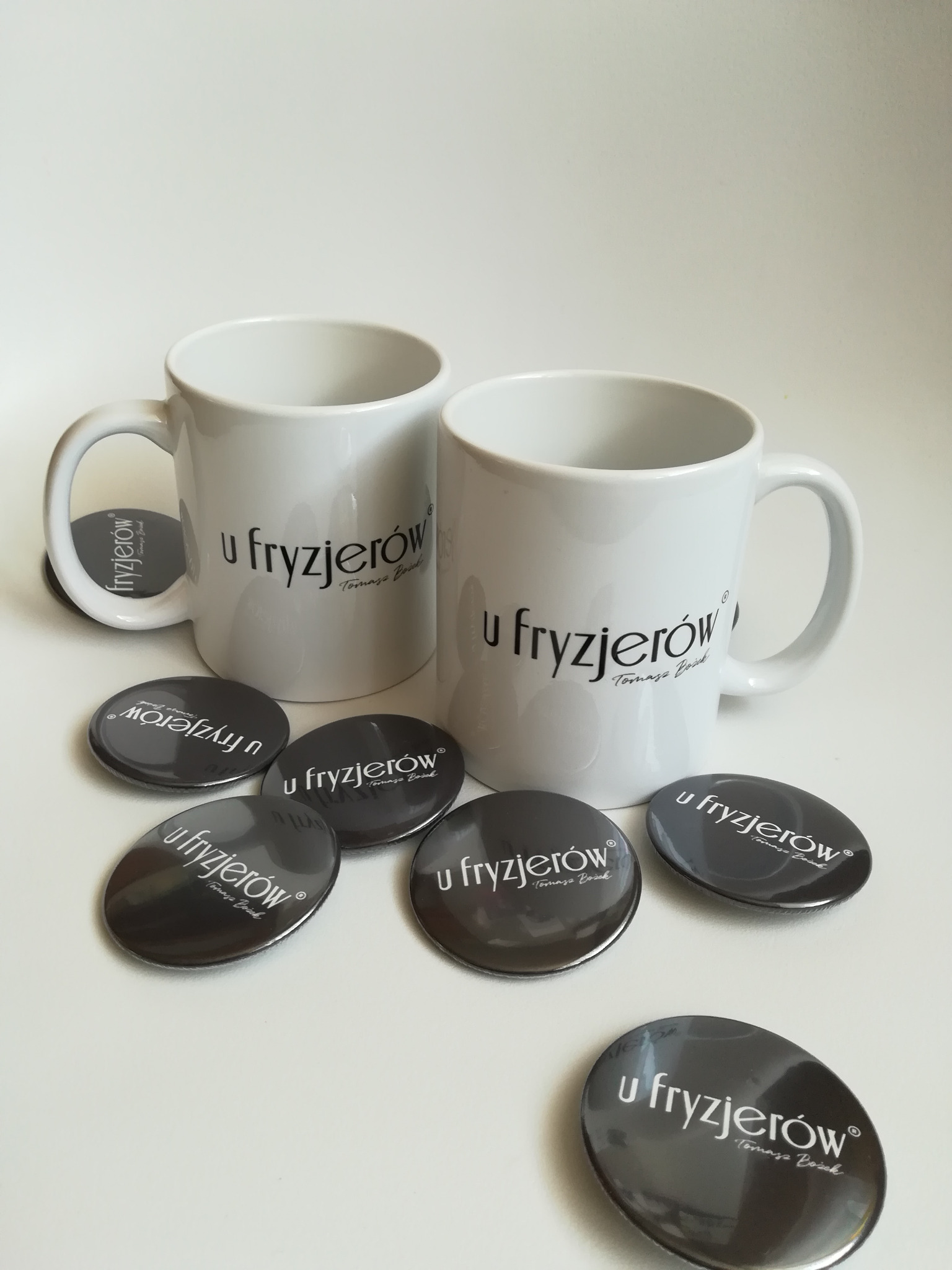 Dwa białe kubki i kilka przypinek z logo 'u fryzjerów Tomasza Bożka' na białym tle.