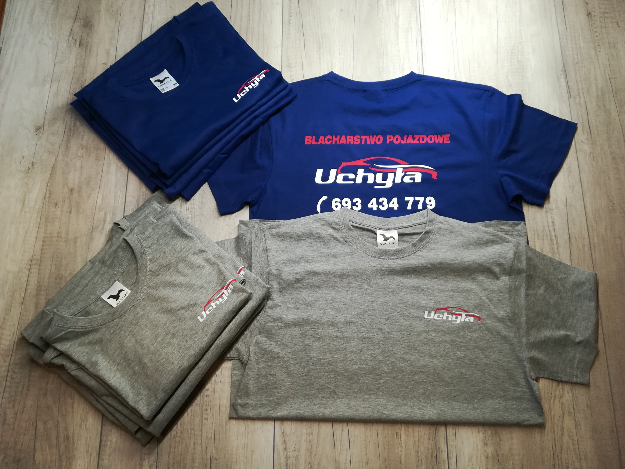 Stos niebieskich i szarych koszulek z nadrukowanym logo 'Uchyla' oraz napisem 'Blacharstwo Pojazdowe' i numerem telefonu, ułożonych na drewnianej powierzchni.