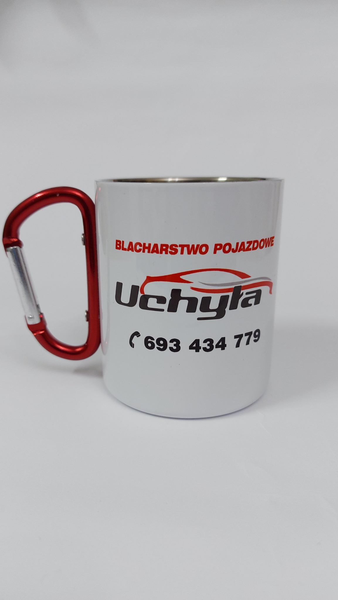 Biały kubek z karabińczykiem jako uchwyt, nadrukiem firmy blacharskiej 'Uchyla' oraz numerem telefonu, na białym tle.