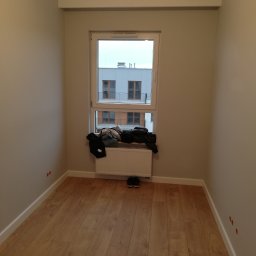 LENDOM Remonty-Wykończenia - Pusty pokój z panelami podłogowymi, świeżo pomalowanymi ścianami w odcieniach beżu, białym grzejnikiem pod oknem, nad którym leżą ubrania, oraz widocznymi otworami na gniazdka elektryczne w ścianie.