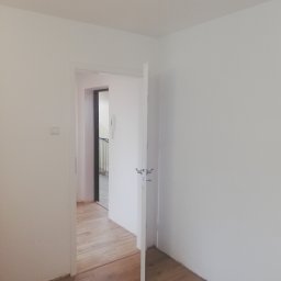 LENDOM Remonty-Wykończenia - Pomalowane na biało wnętrze pokoju z drewnianą podłogą i otwartymi białymi drzwiami, widok na klatkę schodową.