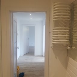 LENDOM Remonty-Wykończenia - Widok na odnowione wnętrze z białymi ścianami, drewnopodobną podłogą i grzejnikiem drabinkowym w łazience, z widocznymi detalami wykończenia.