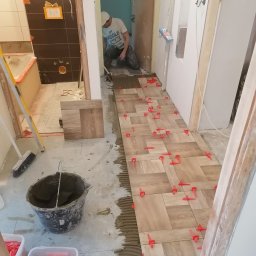 LENDOM Remonty-Wykończenia - Układanie płytek podłogowych w jodełkę, widoczne czerwone klipsy poziomujące, wiadro z zaprawą i płytki na ścianie łazienki w tle.