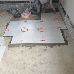 LENDOM Remonty-Wykończenia - Układanie jasnych płytek podłogowych z użyciem krzyżyków dystansowych; widoczny pracownik w trakcie układania, wiadro z zaprawą i narzędzia.