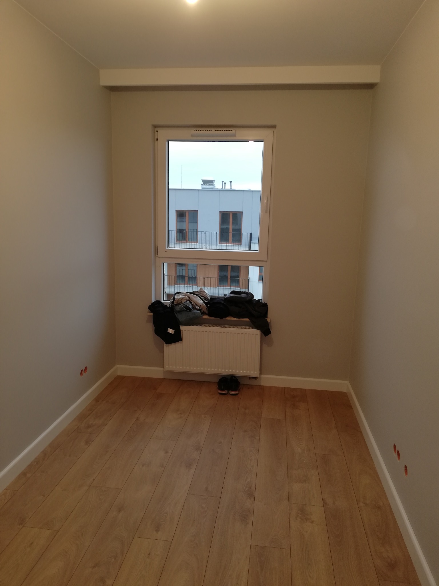 Pusty pokój z panelami podłogowymi, świeżo pomalowanymi ścianami w odcieniach beżu, białym grzejnikiem pod oknem, nad którym leżą ubrania, oraz widocznymi otworami na gniazdka elektryczne w ścianie.