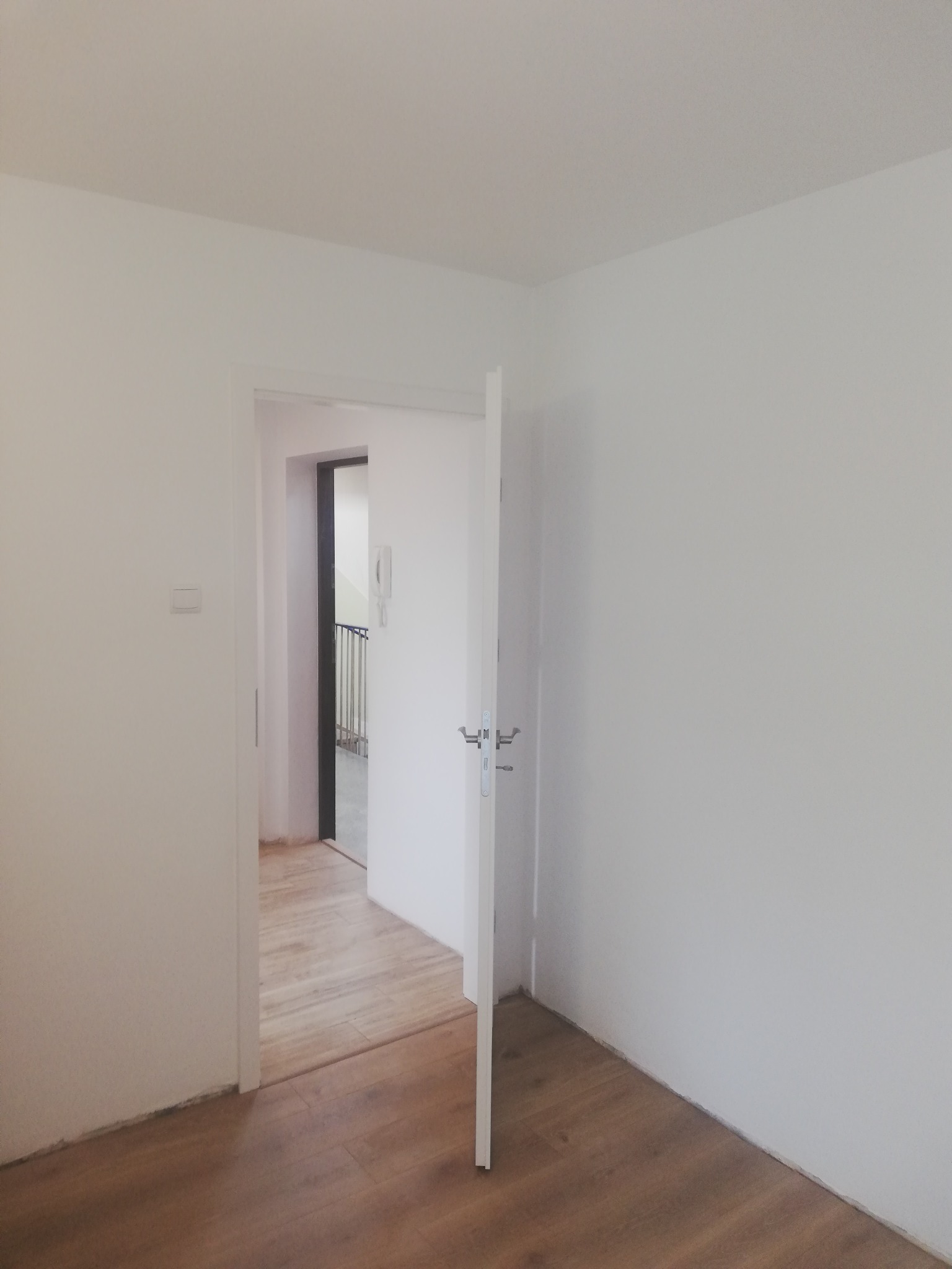 Pomalowane na biało wnętrze pokoju z drewnianą podłogą i otwartymi białymi drzwiami, widok na klatkę schodową.