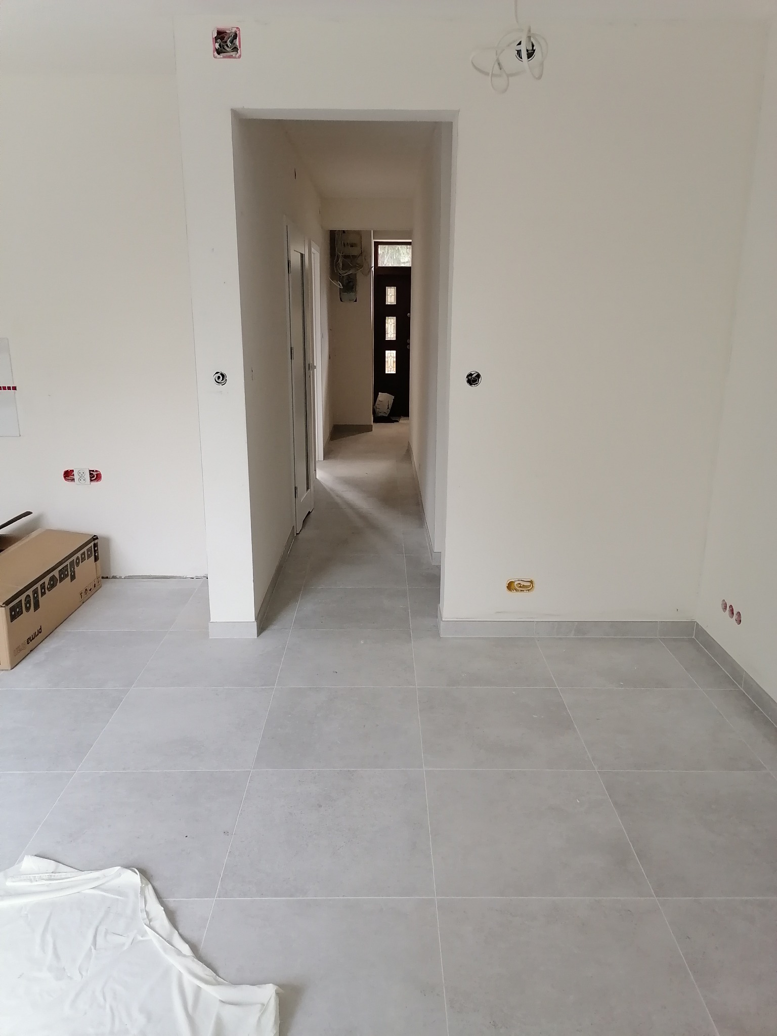 Wnętrze mieszkania po remoncie: świeżo pomalowane białe ściany, szare płytki podłogowe, widoczne gniazdka elektryczne i kartonowe pudło przy ścianie.