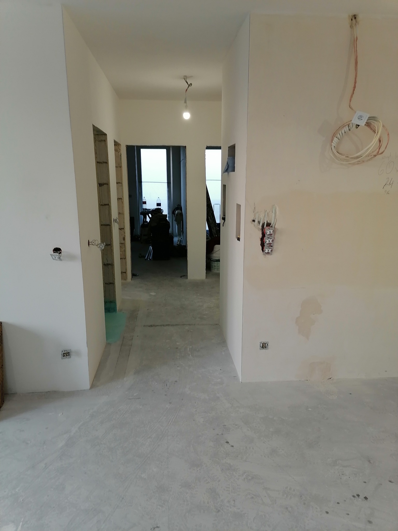 Wnętrze w trakcie prac remontowych: surowe ściany z widocznymi instalacjami elektrycznymi, betonowa podłoga, widok na kolejne pomieszczenia w głębi.