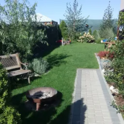 Zadbany ogród w Środzie Wielkopolskiej z równo przystrzyżonym trawnikiem, ścieżką z kostki brukowej, drewnianą ławką, miejscem na ognisko oraz ozdobną drewnianą ścianką z pnącymi roślinami...