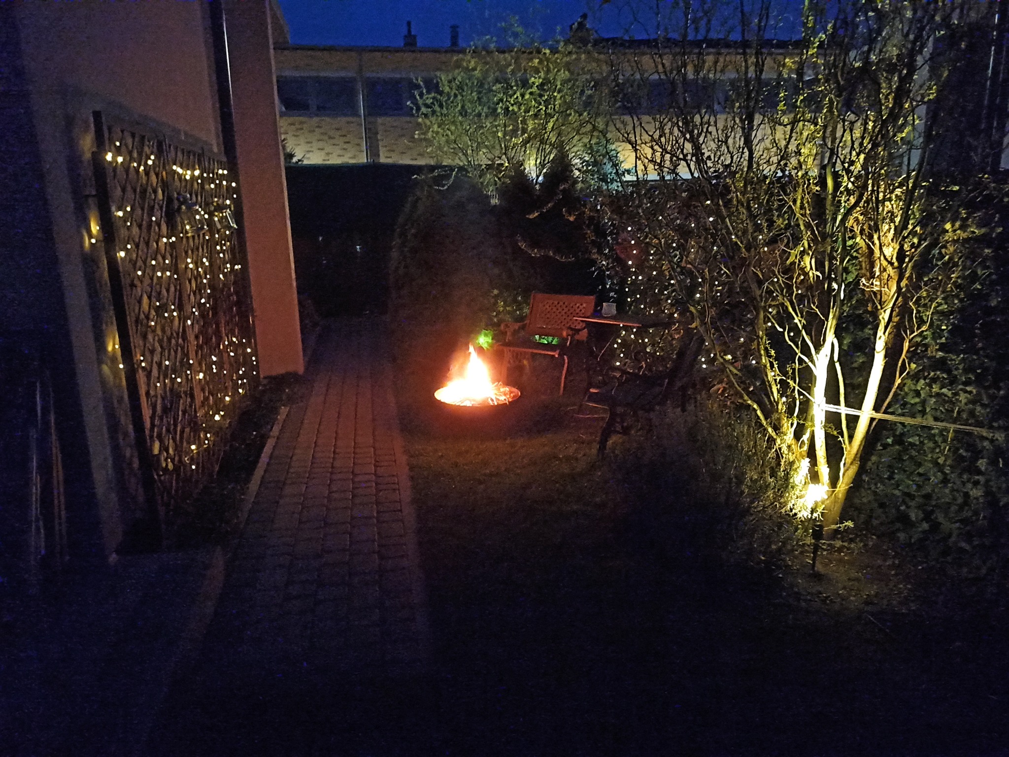 Wieczorny ogród z paleniskiem, oświetlonym drzewem i dekoracyjną ścianką z lampkami, tworzący przytulną atmosferę.