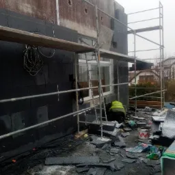 Pracownik w żółtej kamizelce montuje czarne płyty izolacyjne na elewacji budynku, widoczne rusztowania i pozostałości materiału na ziemi.