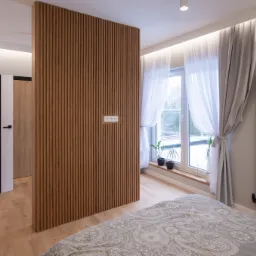 Nowoczesna sypialnia z drewnianą ścianką działową z pionowych listew, gniazdkami elektrycznymi, oknem z białymi zasłonami, czarnym grzejnikiem i fragmentem ł&oacute;żka z szarą pościelą w ornamenty.