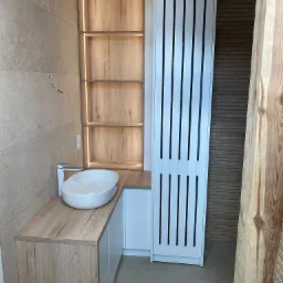 Nowoczesna łazienka z umywalką nablatową na szafce z drewnopodobnym blatem, obok wysoka szafka z białymi lamelami i podświetlana półka wnękowa z drewnianym wykończeniem.