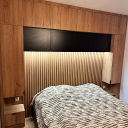 Happy Dreams - Drewniana zabudowa sypialni z szafkami nad łóżkiem i panelem dekoracyjnym z podświetleniem. Widoczne szafki nocne i wzorzysta narzuta na łóżku.