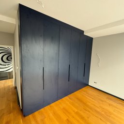 Happy Dreams - Granatowa szafa wnękowa z długimi, czarnymi uchwytami, na tle parkietu i szarej ściany. Widoczne wystające kable ze ściany i sufitu. W tle drzwi z abstrakcyjnym wzorem.