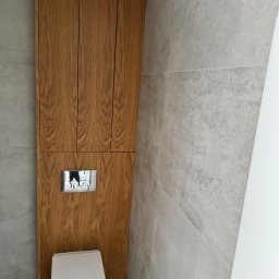 Happy Dreams - Drewniana zabudowa toalety z szafkami wiszącymi, szarymi płytkami i chromowanym przyciskiem spłukiwania. Minimalistyczny design, jasne wnętrze.