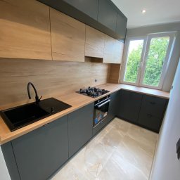 Happy Dreams - Nowoczesna kuchnia na wymiar z drewnianymi frontami szafek górnych i grafitowymi dolnymi. Czarny zlew i bateria, płyta gazowa przy oknie. Marmurowe płytki podłogowe.