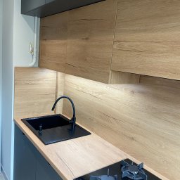 Happy Dreams - Nowoczesna kuchnia z drewnianymi frontami szafek i czarnym blatem, z widocznym zlewem i płytą grzewczą. Oświetlenie LED pod szafkami górnymi.