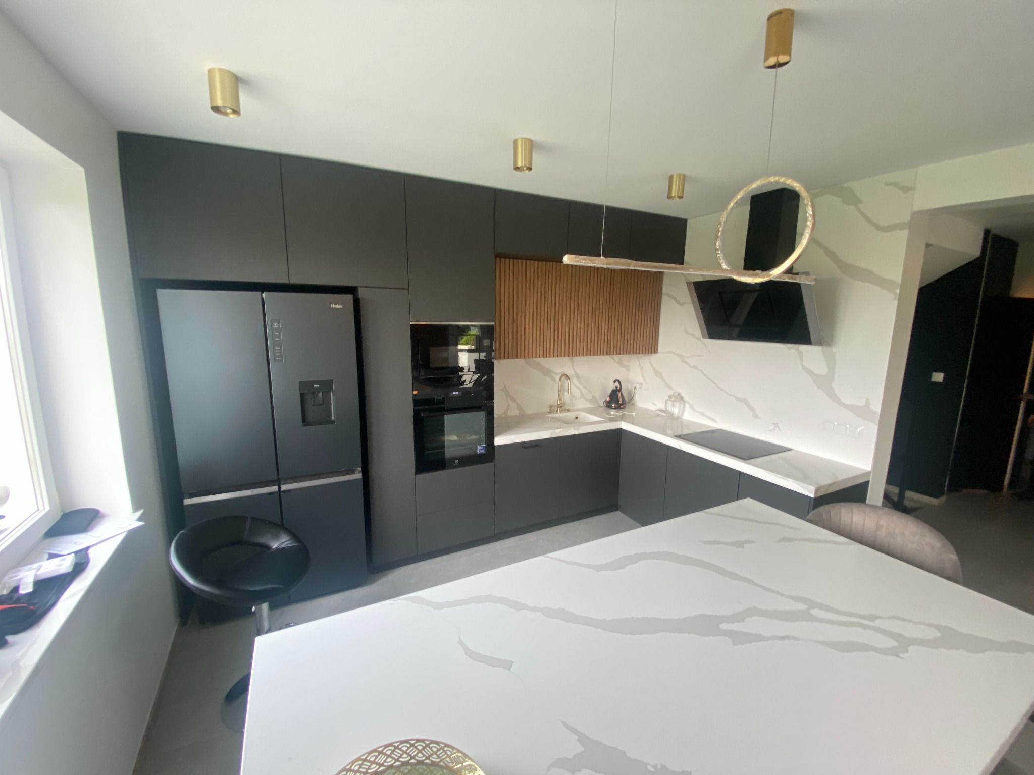 Nowoczesna kuchnia z ciemnymi szafkami, marmurowym blatem i złotymi akcentami oświetleniowymi, widok na wyspę kuchenną i okno.