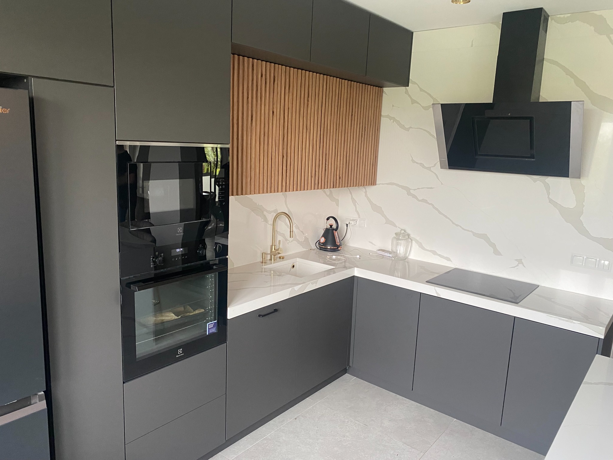 Nowoczesna kuchnia w odcieniach szarości z drewnianym panelem dekoracyjnym, białym blatem z marmurowym wzorem i złotą armaturą, czajnik elektryczny na blacie, piekarnik zabudowany.