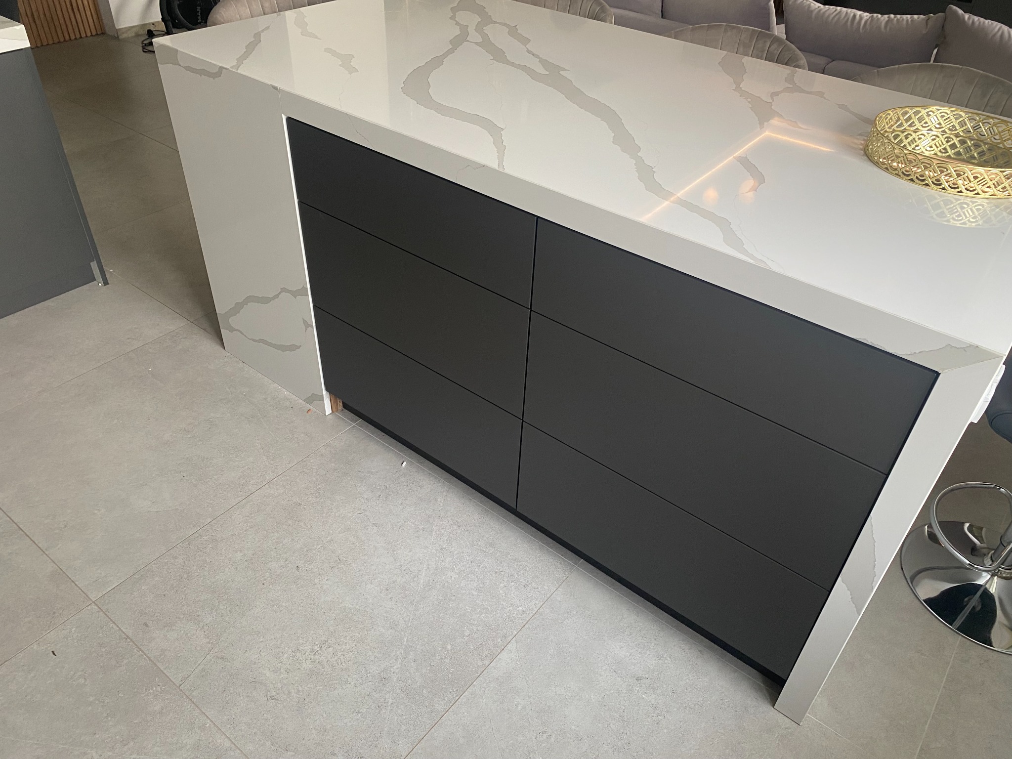 Wyspa kuchenna z blatem imitującym marmur i szufladami w kolorze antracytowym, widoczny fragment salonu w tle.