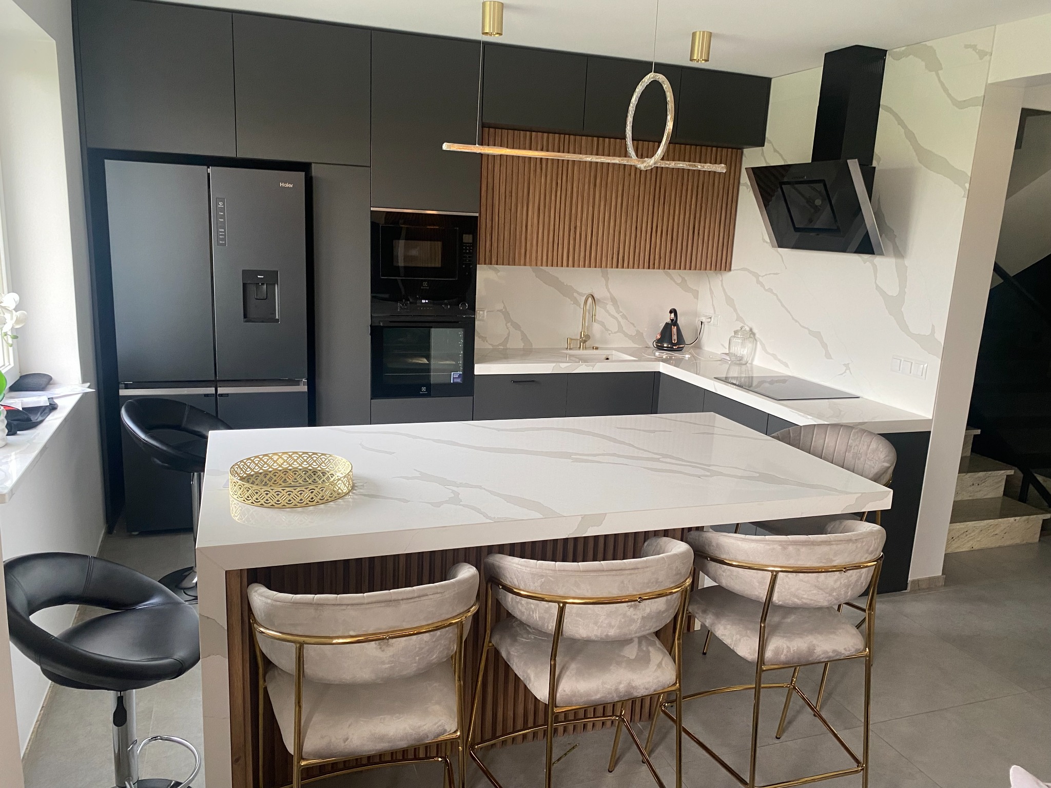Nowoczesna kuchnia z czarnymi szafkami, wyspą z blatem imitującym marmur, złotymi akcentami oświetlenia i krzesłami barowymi z welurową tapicerką