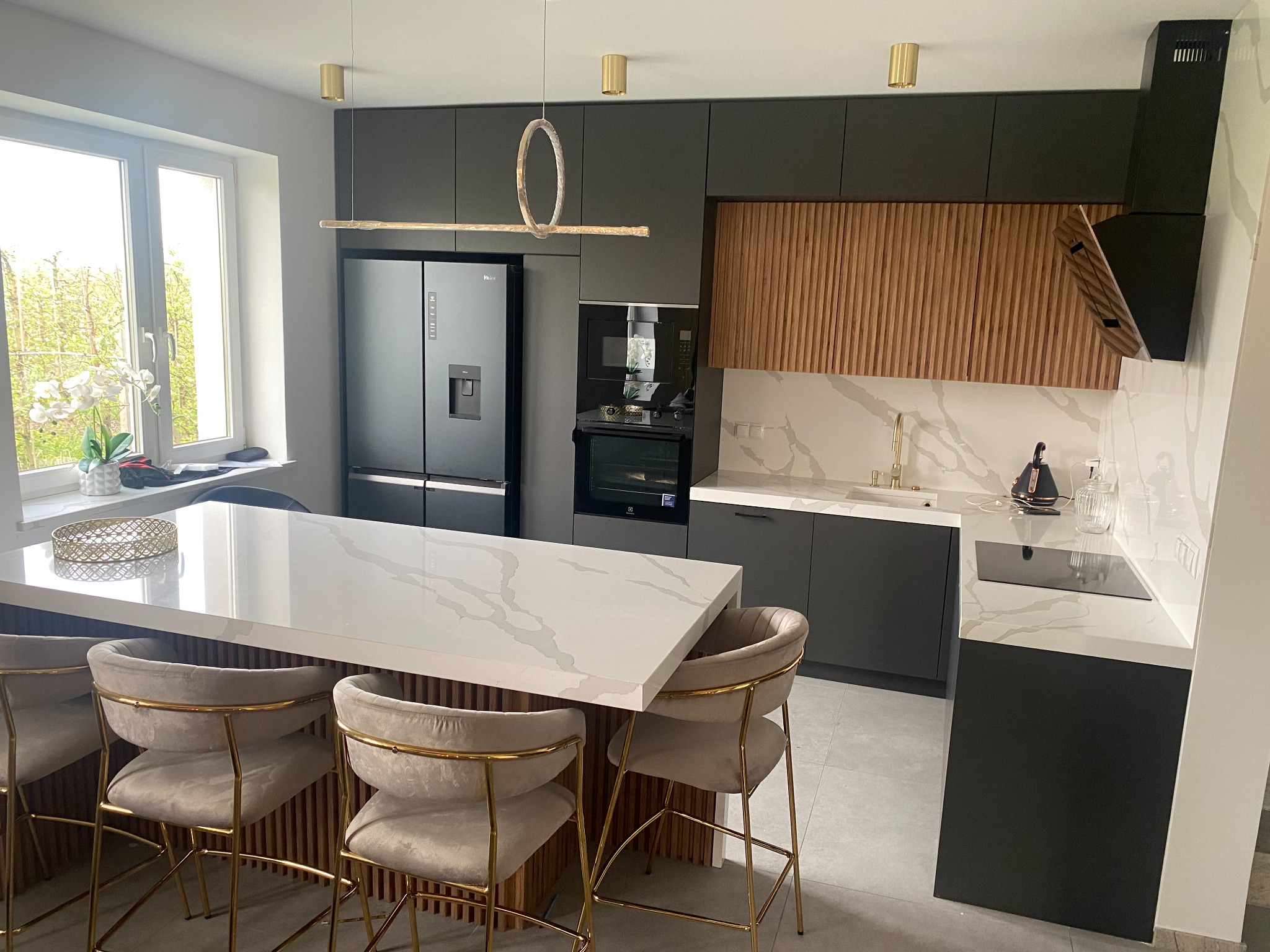 Nowoczesna kuchnia z szarymi szafkami, drewnianymi akcentami na ścianie i wyspą z białym blatem imitującym marmur, oświetlona złotymi lampami wiszącymi. Krzesła barowe z beżową tapicerką i złotymi...