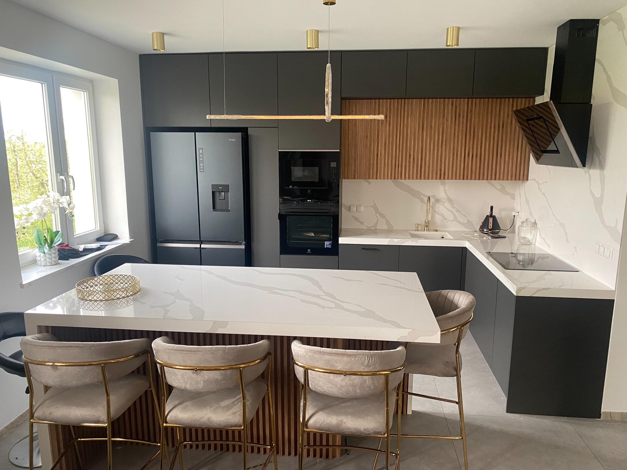 Nowoczesna kuchnia z ciemnoszarymi szafkami, wyspą z blatem imitującym marmur, drewnianymi akcentami na ścianie i wyspie, oraz złotymi detalami oświetlenia i krzesel barowych.