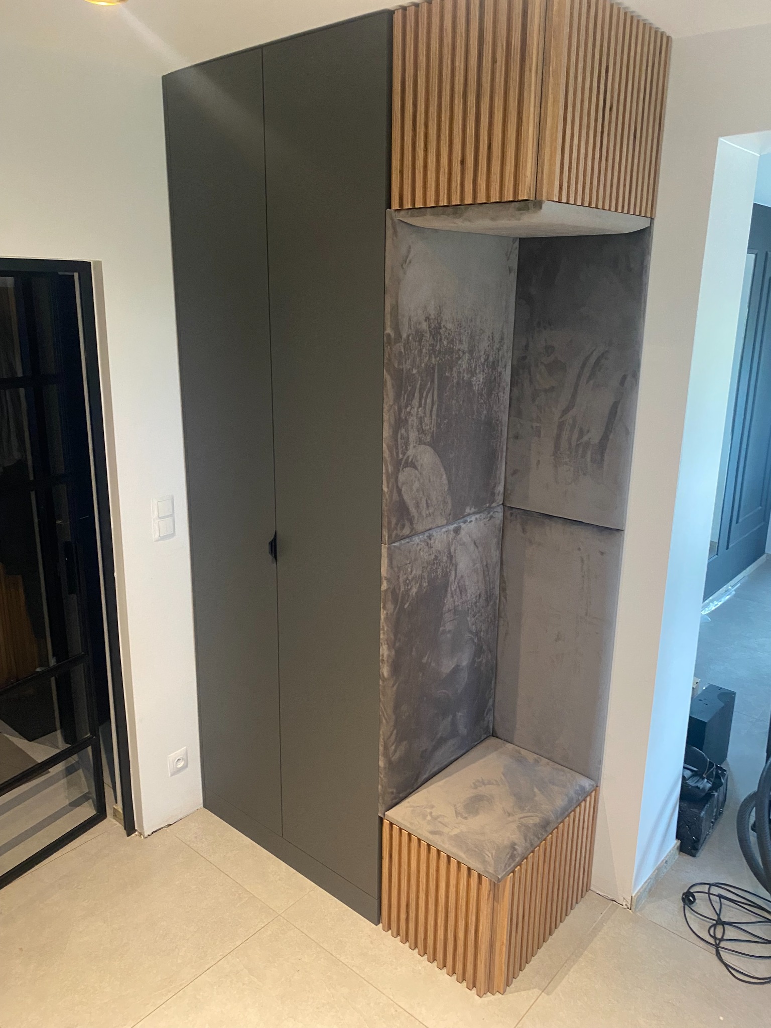 Szara szafa z siedziskiem wnękowym wykończonym drewnianymi lamelami i tapicerowanymi panelami ściennymi w odcieniach szarości, widok na fragment podłogi z płytek i drzwi wejściowe.